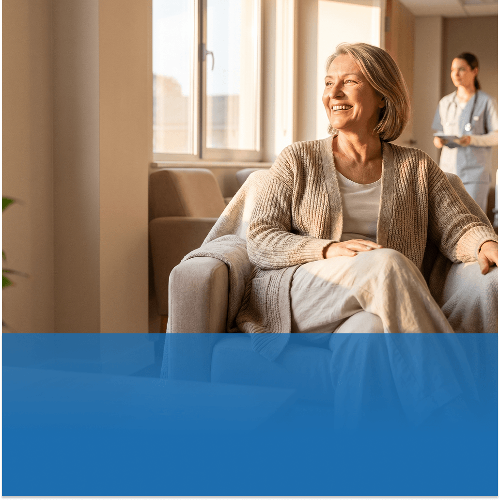 Smiling older woman sits on a beige sofa in a bright waiting area, with a healthcare worker in scrubs in the background.